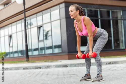 Active lifestyle concept. Fit woman in sportswear trains muscles with dumbbells in hands outdoors