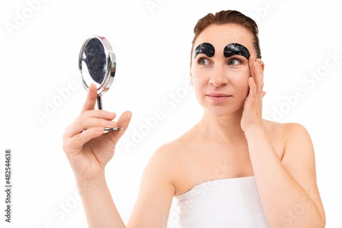 young woman with cosmetic patches on her eyebrows looks in the mirror on a white background