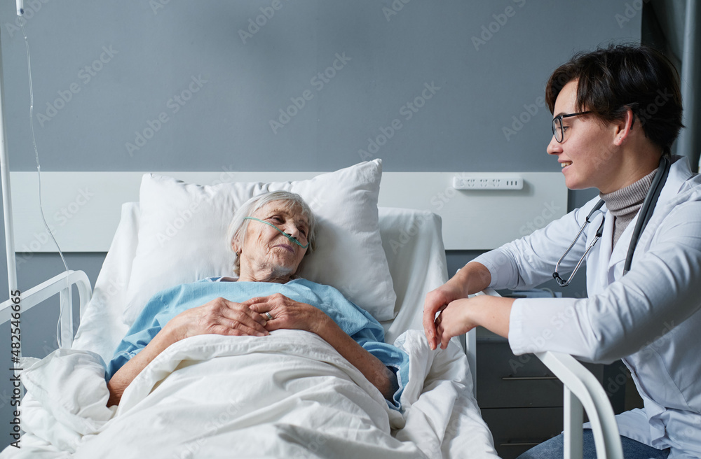 Sick senior woman resting on the bed and talking to the nurse who caring about her at hospital - obrazy, fototapety, plakaty 