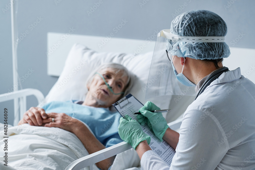 Obraz premium Rear view of young doctor in protective wear writing prescription for senior woman while she lying on bed at the ward