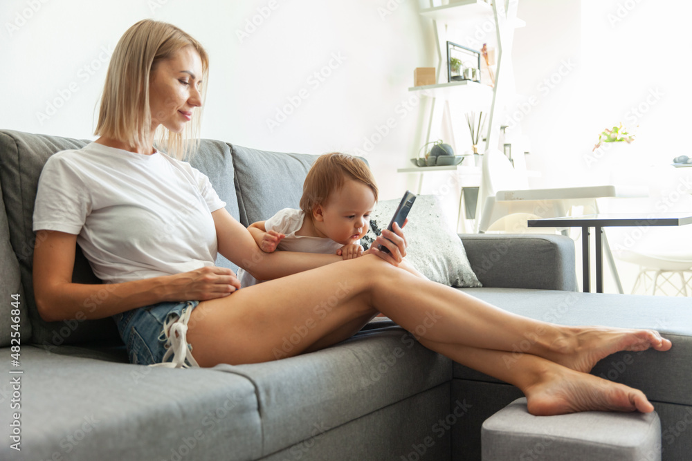 Cute young mother with her little daughter uses a smart phone while sitting on the couch at home Cute young mother with her little daughter uses a smart phone while sitting on the couch at home