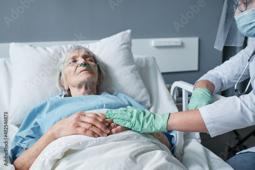 Doctor in protective gloves holding hands of senior woman while she lying on bed at ward, doctor caring about her during disease