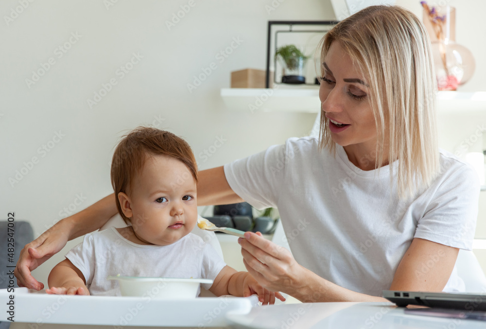 A caring mother feeds her baby from a spoon while sitting at a table A caring mother feeds her baby from a spoon while sitting at a table