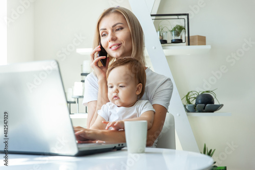 Business mom with little daughter in her arms uses a laptop and talking on phone while sitting at a table at home