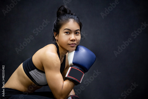 Attractive Asian boxer woman sitting and posing with blue boxing gloves in fitness gym. Female boxing class and martial art concept