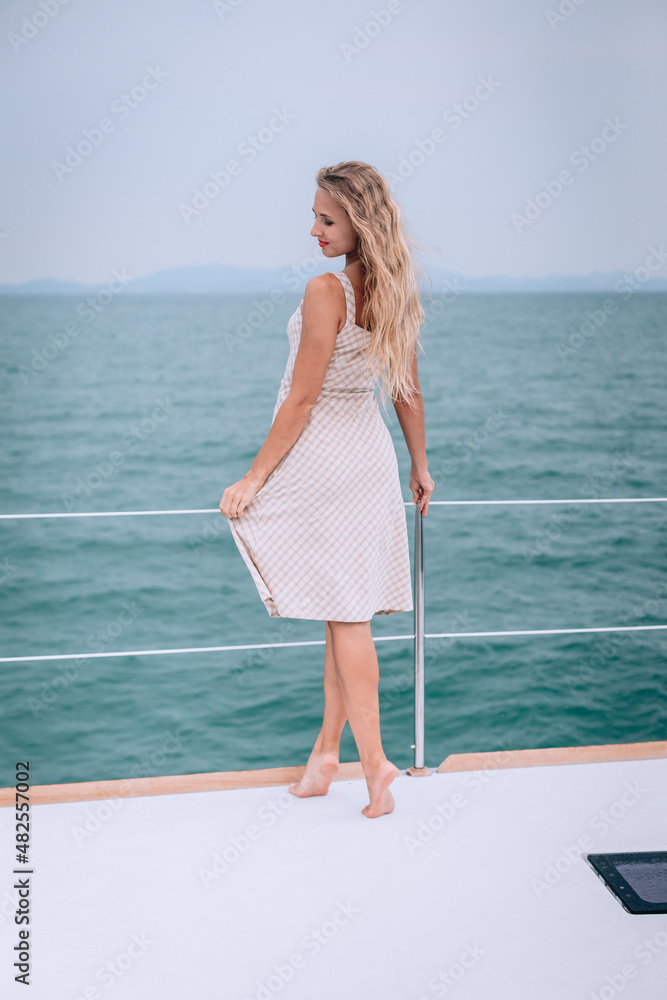 Outdoor photography. A gentle, wonderful young skinny girl with long hair poses for the camera on a white yacht in the open turquoise sea in a beige checkered dress. A face with slightly lowered eyes.