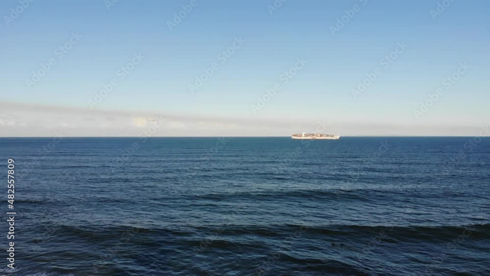 A large cargo ship sails through large, open waters off the coast to a nearby beach town under a baby blue sky that harbours low hanging fluffy clouds A large cargo ship sails through large, open waters off the coast to a nearby beach town under a baby blue sky that harbours low hanging fluffy clouds