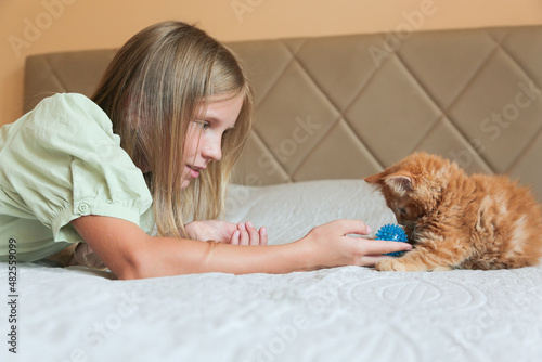 Happy girl playing with her ginger cat on a bed. Blue-eyed girl and kitten. Child and pet.