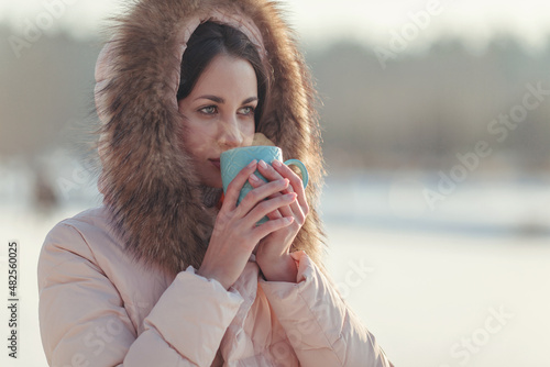 Сlose-up photo taken in winter. A beautiful young green-eyed girl with dark hair and a pink down jacket with fur is holding a blue cup of tea. The desire to get comfort and warmth in the cold period.