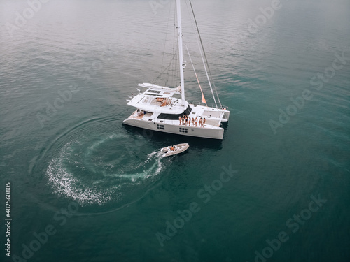 A wonderful aerial view photo of a white yacht with several young awesome girls in swimsuits looking at a guy on a white motorboat. Drive up to the yacht. The concept of expensive vacation.