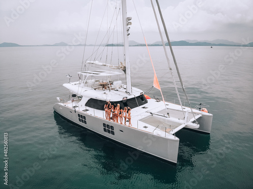 A photo created by drone. A lovely young women in a beautiful swimsuit pose on a white expensive luxury yacht in the bay open sea.Demonstration of luxury life. The concept of women s beauty.