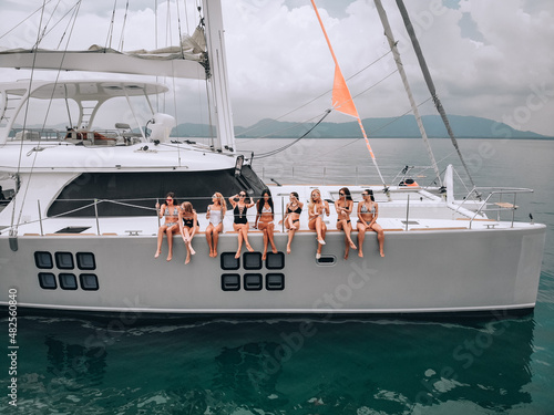 A photo-session of a cute young girls in swimsuits and sunglasses on a white beautiful yacht in the open turquoise sea. Behind the bigisland. The concept of a holiday in the tropics. photo