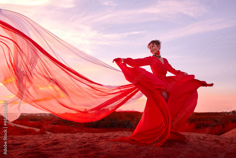 Beautiful young woman or girl with curly hair and in red dress with a light flying fabric on the sand on sunny day with blue sky in the background. Model or dancer posing in photo shoot on sand dunes