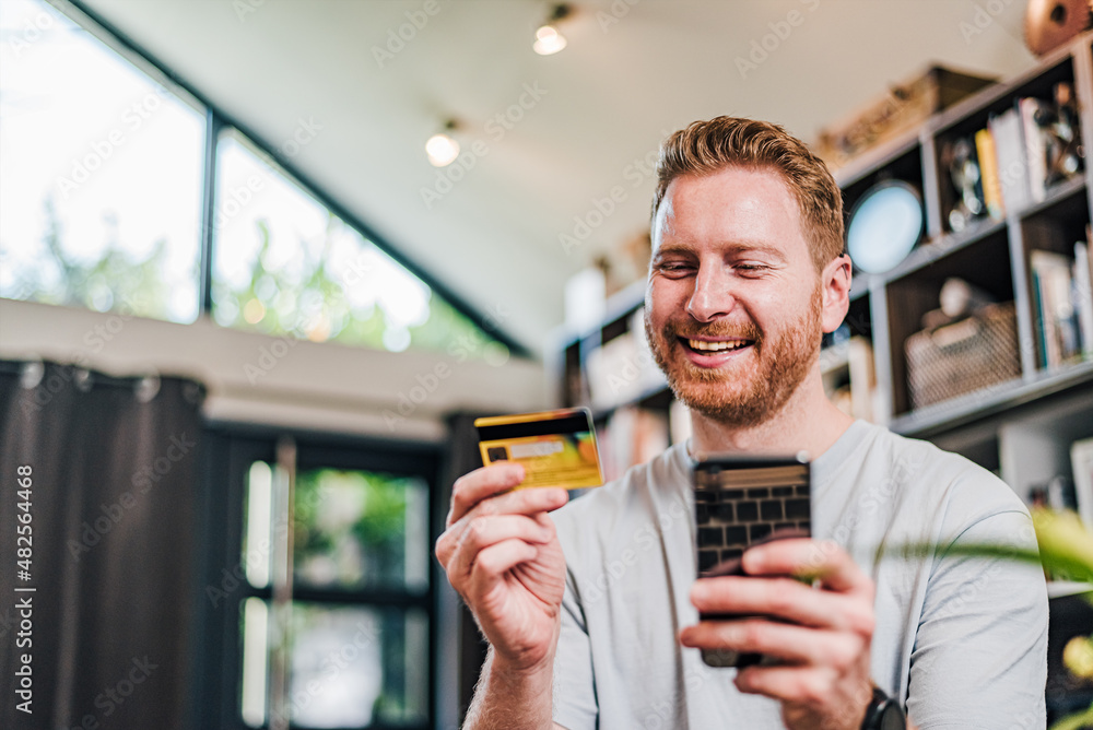 Low angle shop of handsome young adult smiling man holding credit card and using phone doing online payment, purchasing and shopping. Businessman using hand held devices from home office
