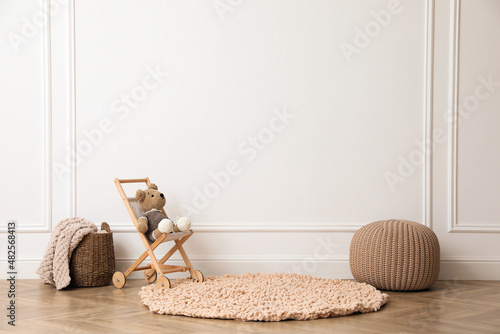 Toy stroller with bear, pouf and wicker basket near white wall in child room. Interior design