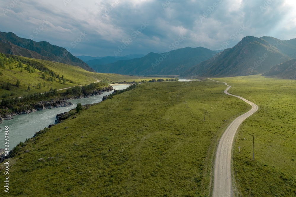 Summer view of a mountain range with a green lawn, a narrow road and a flowing river under blue sky. Summer view of a mountain range with a green lawn, a narrow road and a flowing river under blue sky.