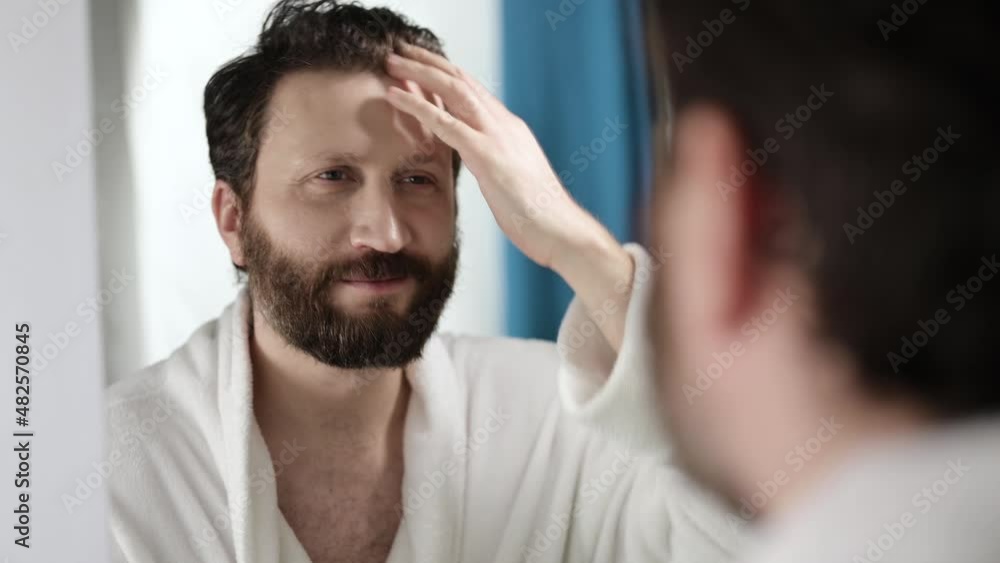 A handsome bearded man looks at himself in mirror after shower. Admire herself, straighten her hair. A man is getting ready for working day. A handsome bearded man looks at himself in mirror after shower. Admire herself, straighten her hair. A man is getting ready for working day.
