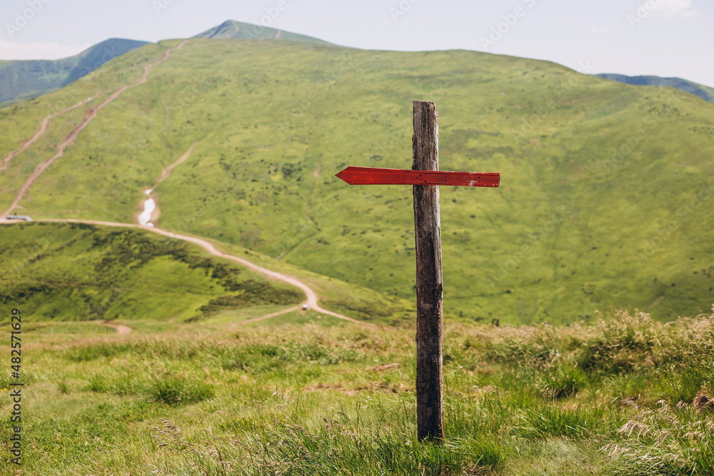 One wooden red signpost in the summer mountain with copyspace. Conceptual leading indicator. Old Arrow on the wooden signpost pointing to a direction. One wooden red signpost in the summer mountain with copyspace. Conceptual leading indicator. Old Arrow on the wooden signpost pointing to a direction.