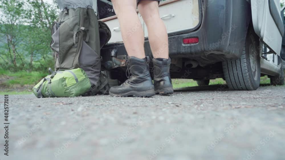 Cropped Image Of A Male Camper Wearing Black Leather Boots Organizing Stuff In A Backpack Outside Rear Compartment Of A Van. medium shot Cropped Image Of A Male Camper Wearing Black Leather Boots Organizing Stuff In A Backpack Outside Rear Compartment Of A Van. medium shot