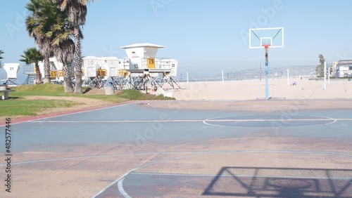 Palm trees and basketball sport field or court on beach, California coast, USA. Streetball playground on shore and lifeguard stand, tower ot station. Mission beach, San Diego. Hoop, backboard and sky.