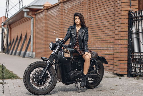 Attractive leggy slender model wearing leather jacket and dress sitting on a black retro motorcycle outside against a backdrop of brick walls and streets. City concept © Semachkovsky 