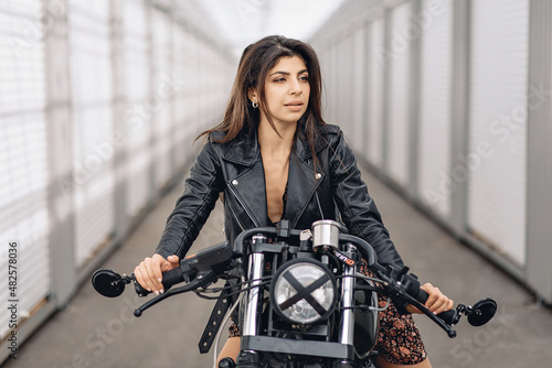 Portrait of a lovely young fit dark-haired female model in a black leather jacket sitting on a black retro motorcycle and looking straight into the camera against a white background. Sexuality concept