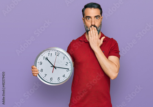 Middle aged man with beard holding big clock covering mouth with hand, shocked and afraid for mistake. surprised expression