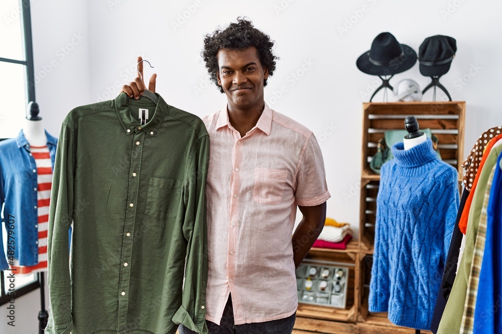 African man with curly hair holding shirt from clothing rack at retail shop looking positive and happy standing and smiling with a confident smile showing teeth