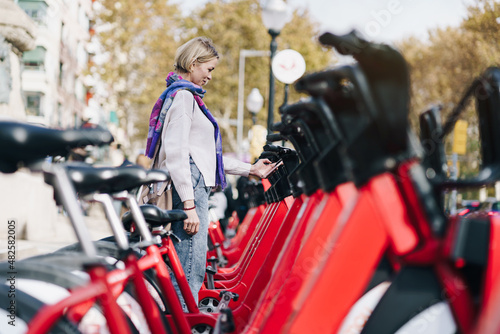 Blond woman taking a red bicycle in a bike rental station in the city with smartphone sharing app - Environment sustainable transport