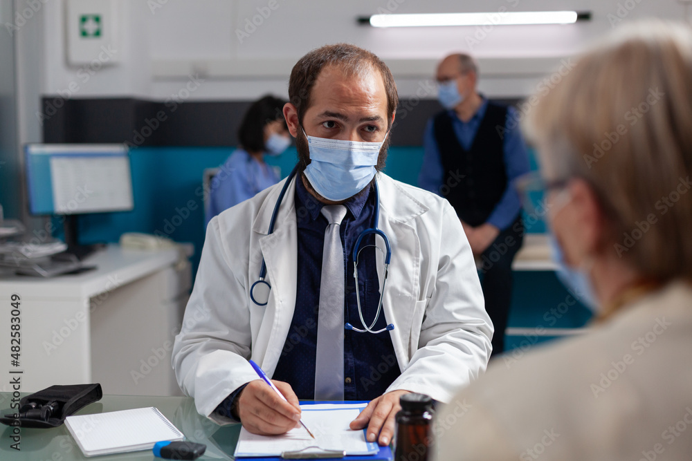 Medic with face mask taking notes at consultation appointment, talking to senior woman in office. Specialist having conversation about medicine and health care with patient during pandemic. Medic with face mask taking notes at consultation appointment, talking to senior woman in office. Specialist having conversation about medicine and health care with patient during pandemic.