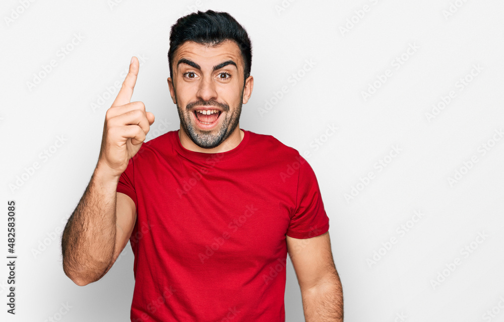 Hispanic man with beard wearing casual red t shirt pointing finger up with successful idea. exited and happy. number one. Hispanic man with beard wearing casual red t shirt pointing finger up with successful idea. exited and happy. number one.