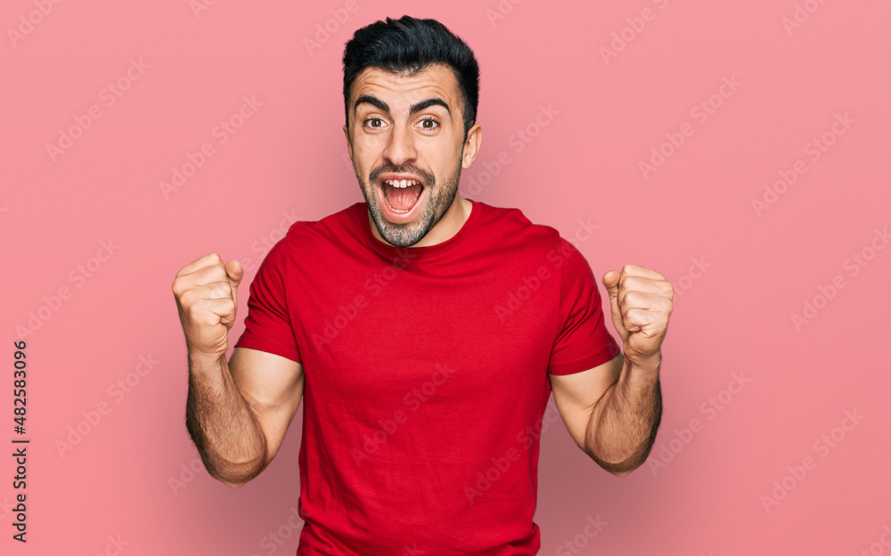 Hispanic man with beard wearing casual red t shirt celebrating surprised and amazed for success with arms raised and open eyes. winner concept. Hispanic man with beard wearing casual red t shirt celebrating surprised and amazed for success with arms raised and open eyes. winner concept.