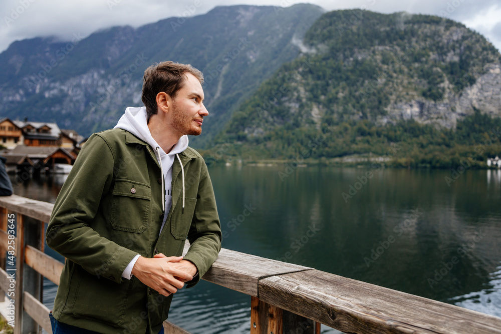 Hallstatt, Austria: Portrait of beautiful romantic young man tourist smile, natural light, boy in green jacket, scenic picturesque town street, mountain village near lake, Alps at summer day