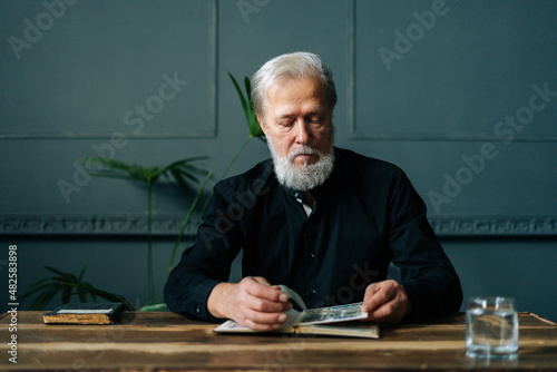 Portrait of serious gray-haired senior adult man looking postage stamps at collection book sitting at wooden table. Focused mature aged man examining album with old stamps philately at home. photo