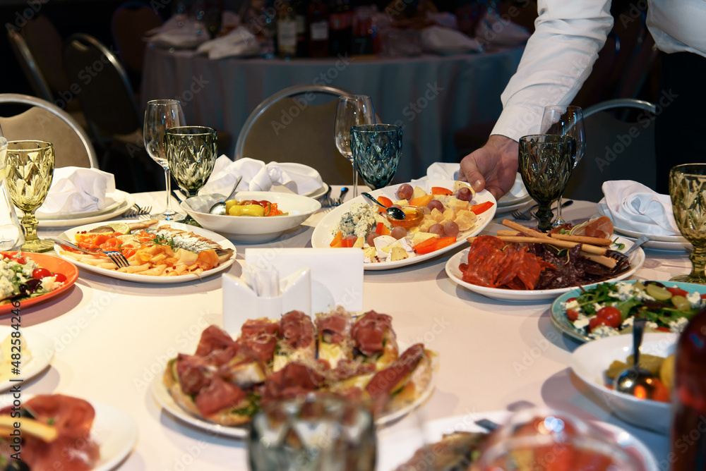 Table is set with variety of gourmet dishes and appetizers for banquet. Waiter's hand putting plate on table. Selective focus. Table is set with variety of gourmet dishes and appetizers for banquet. Waiter's hand putting plate on table. Selective focus.