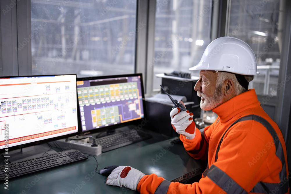 Industrial worker in safety equipment sitting in factory control room monitoring production process and talking on radio communication. Industrial worker in safety equipment sitting in factory control room monitoring production process and talking on radio communication.
