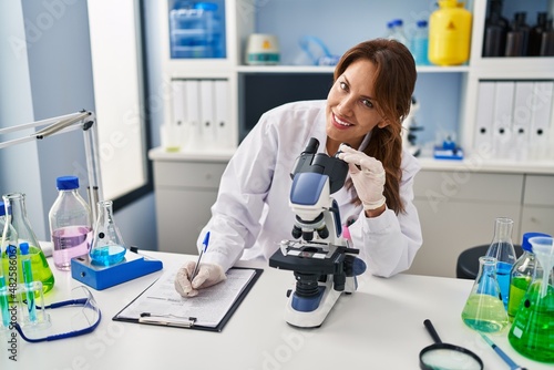 Young latin woman wearing scientist uniform using microscope writing on clipboard at laboratory