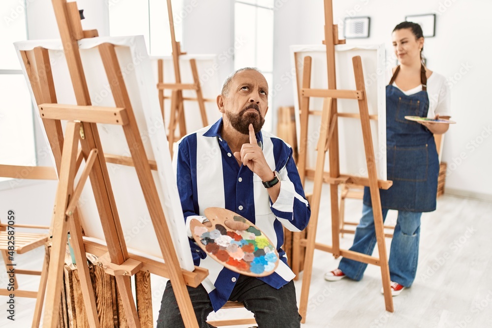Senior artist man at art studio thinking concentrated about doubt with finger on chin and looking up wondering Senior artist man at art studio thinking concentrated about doubt with finger on chin and looking up wondering
