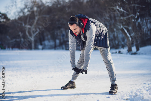 Adult man is exercising in park in wintertime. He is stretching his body before jogging.