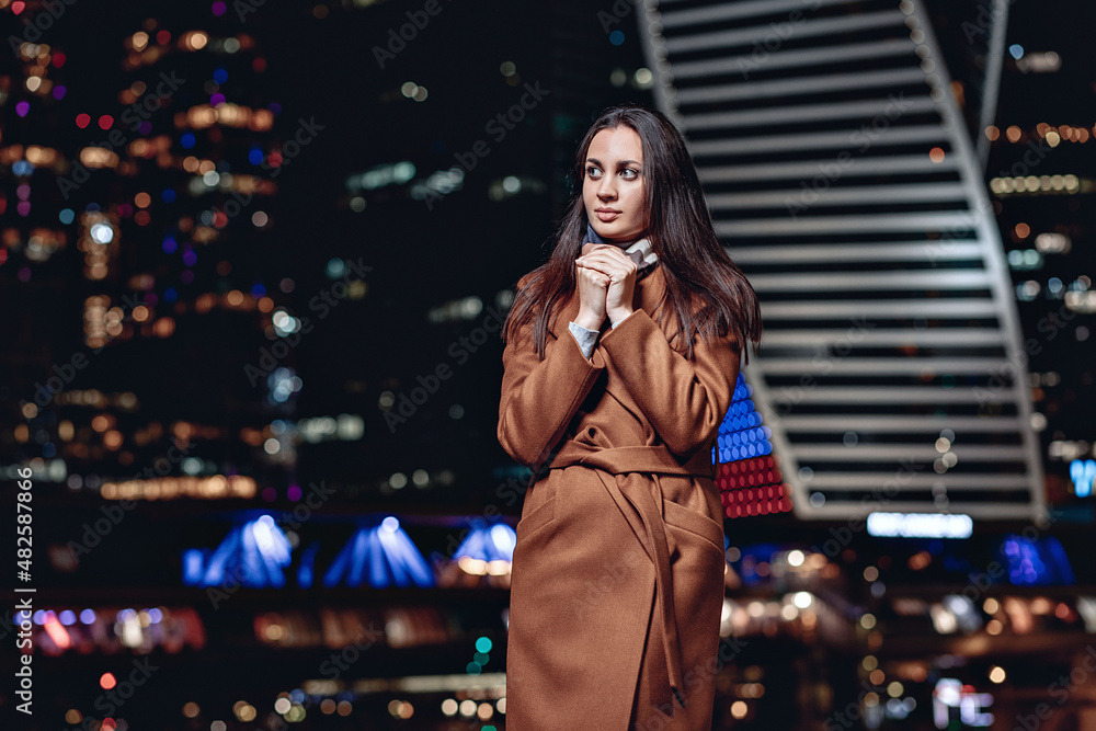Portrait of a cute modest beautiful girl wearing a coat and a scarf and posing against the backdrop of the night Moscow city. Bright city lights concept