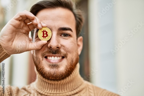 Tablou canvas Young caucasian man smiling happy holding bitcoin over eye at the city