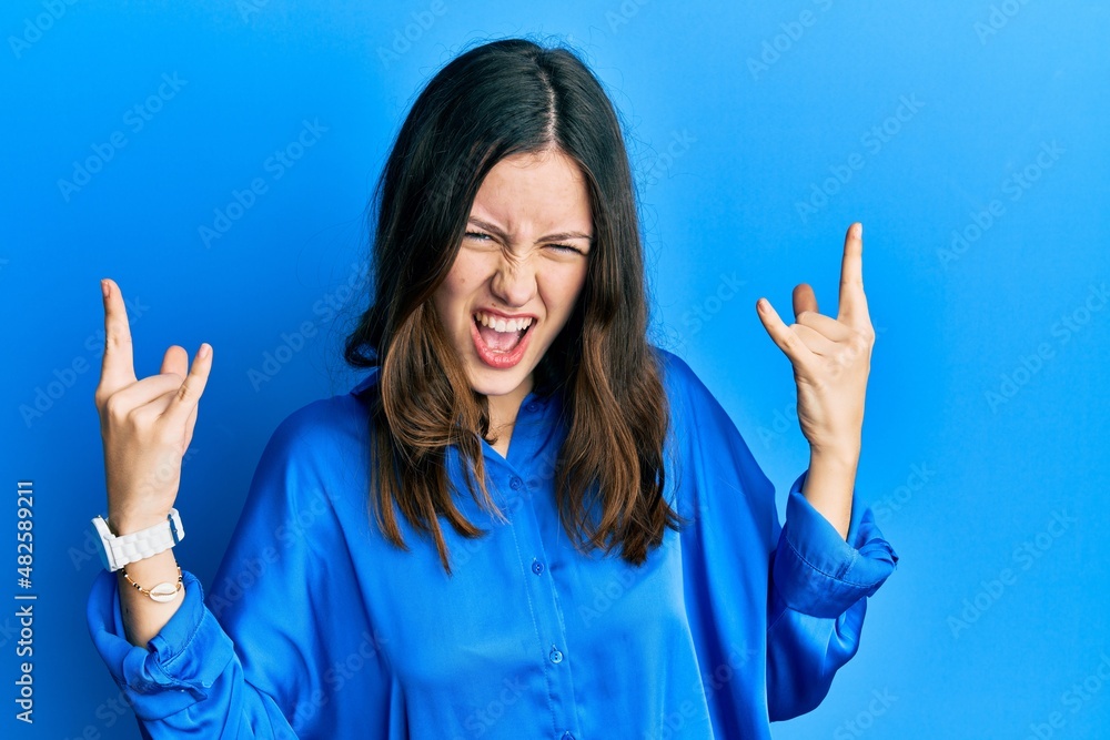 Young brunette woman wearing casual blue shirt shouting with crazy expression doing rock symbol with hands up. music star. heavy concept. Young brunette woman wearing casual blue shirt shouting with crazy expression doing rock symbol with hands up. music star. heavy concept.