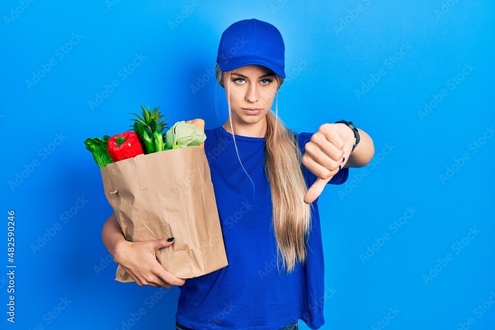 Young caucasian woman wearing courier uniform with groceries from supermarket looking unhappy and angry showing rejection and negative with thumbs down gesture. bad expression. Young caucasian woman wearing courier uniform with groceries from supermarket looking unhappy and angry showing rejection and negative with thumbs down gesture. bad expression.