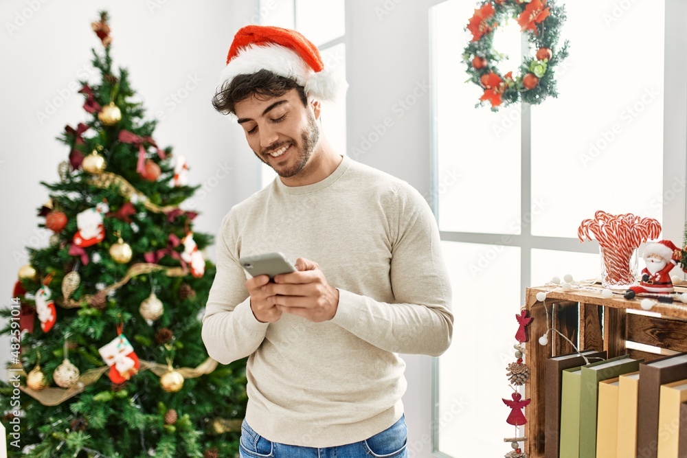 © Krakenimages.com - Young hispanic man wearing christmas hat using smartphone at home.