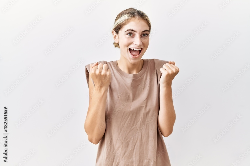 Young blonde woman standing over isolated background celebrating surprised and amazed for success with arms raised and open eyes. winner concept. Young blonde woman standing over isolated background celebrating surprised and amazed for success with arms raised and open eyes. winner concept.