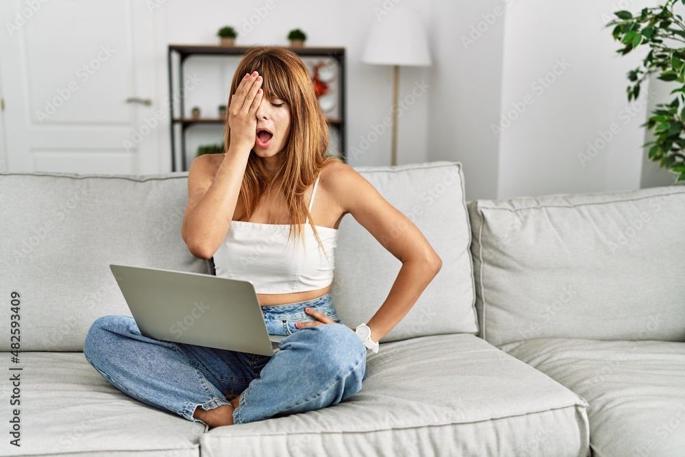 Hispanic woman sitting on the sofa at home using laptop yawning tired covering half face, eye and mouth with hand. face hurts in pain.
