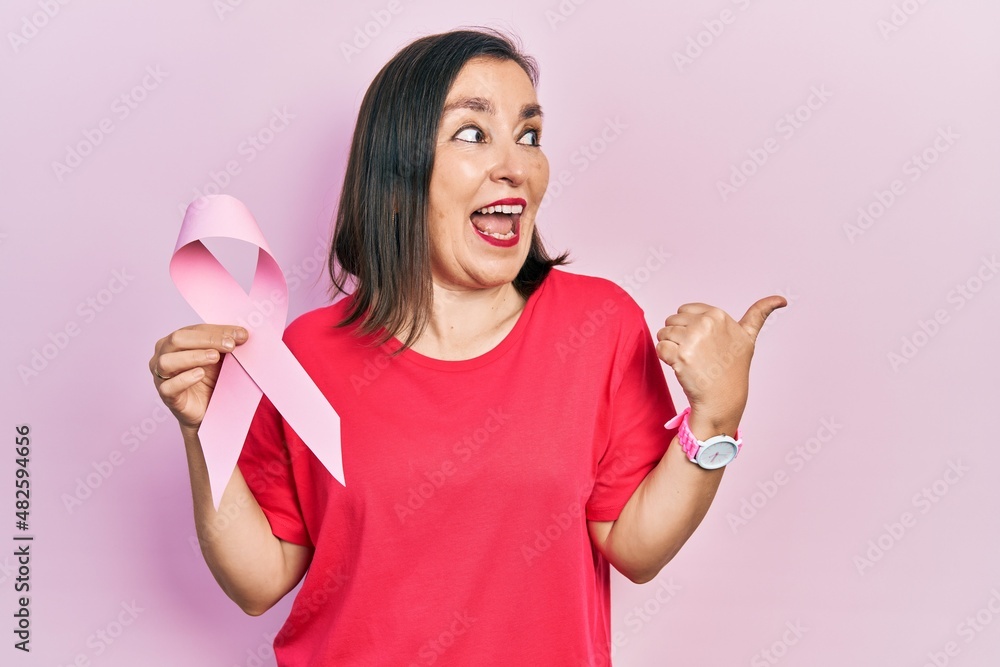 Middle age hispanic woman holding pink cancer ribbon pointing thumb up to the side smiling happy with open mouth Middle age hispanic woman holding pink cancer ribbon pointing thumb up to the side smiling happy with open mouth
