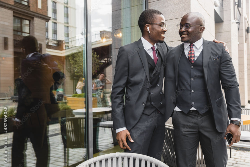 Two dark-skinned African American businessmen friends in suits hug and rejoice at a meeting on the street near a cafe Two dark-skinned African American businessmen friends in suits hug and rejoice at a meeting on the street near a cafe