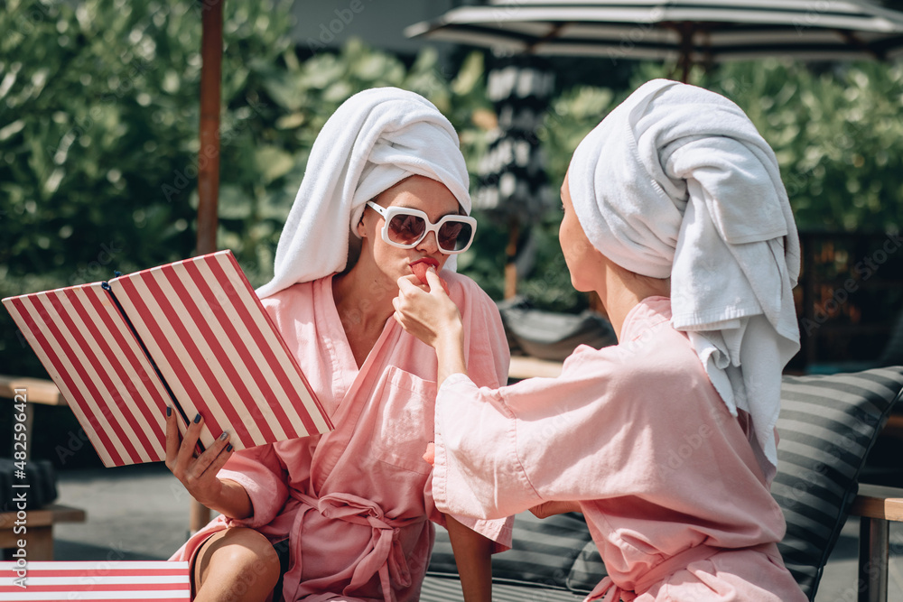 Portrait of two sexy charming ladies wearing identical bathrobes and towels on their heads are spending time together. Nice girl feeding her friend a tasty watermelon. Friendship concept