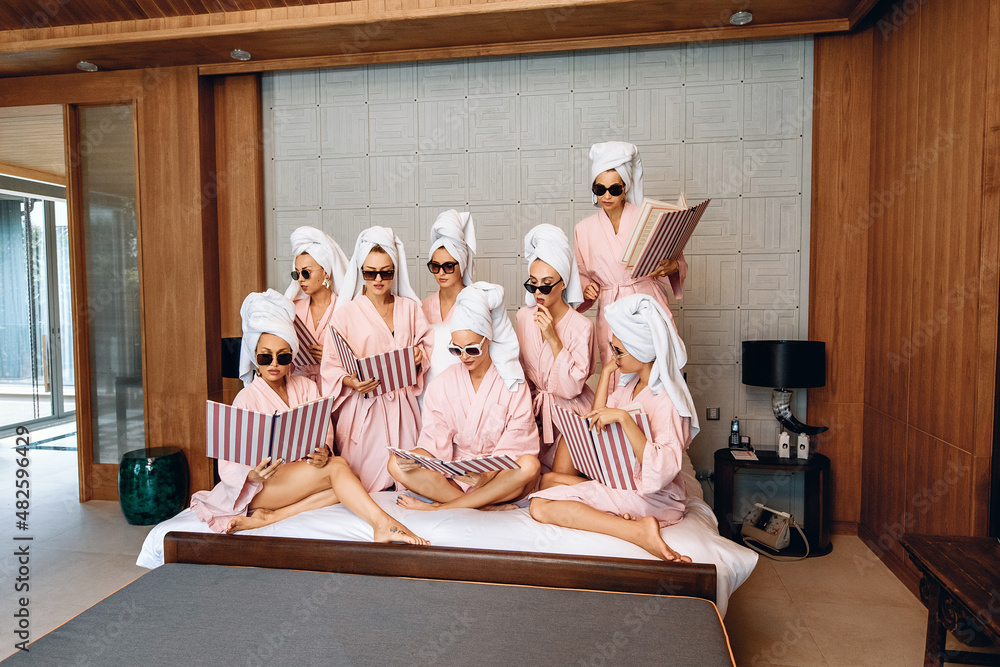 Group of adorable focused female models in the same clothes wearing bathrobes, a towel on their heads and sunglasses and posing with a menu in their hands indoor. Self-care concept